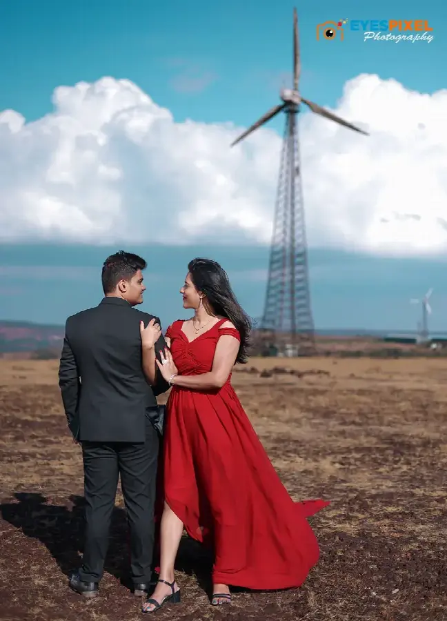 Couple on open grassland below a wind turbine, she in a flowing red gown